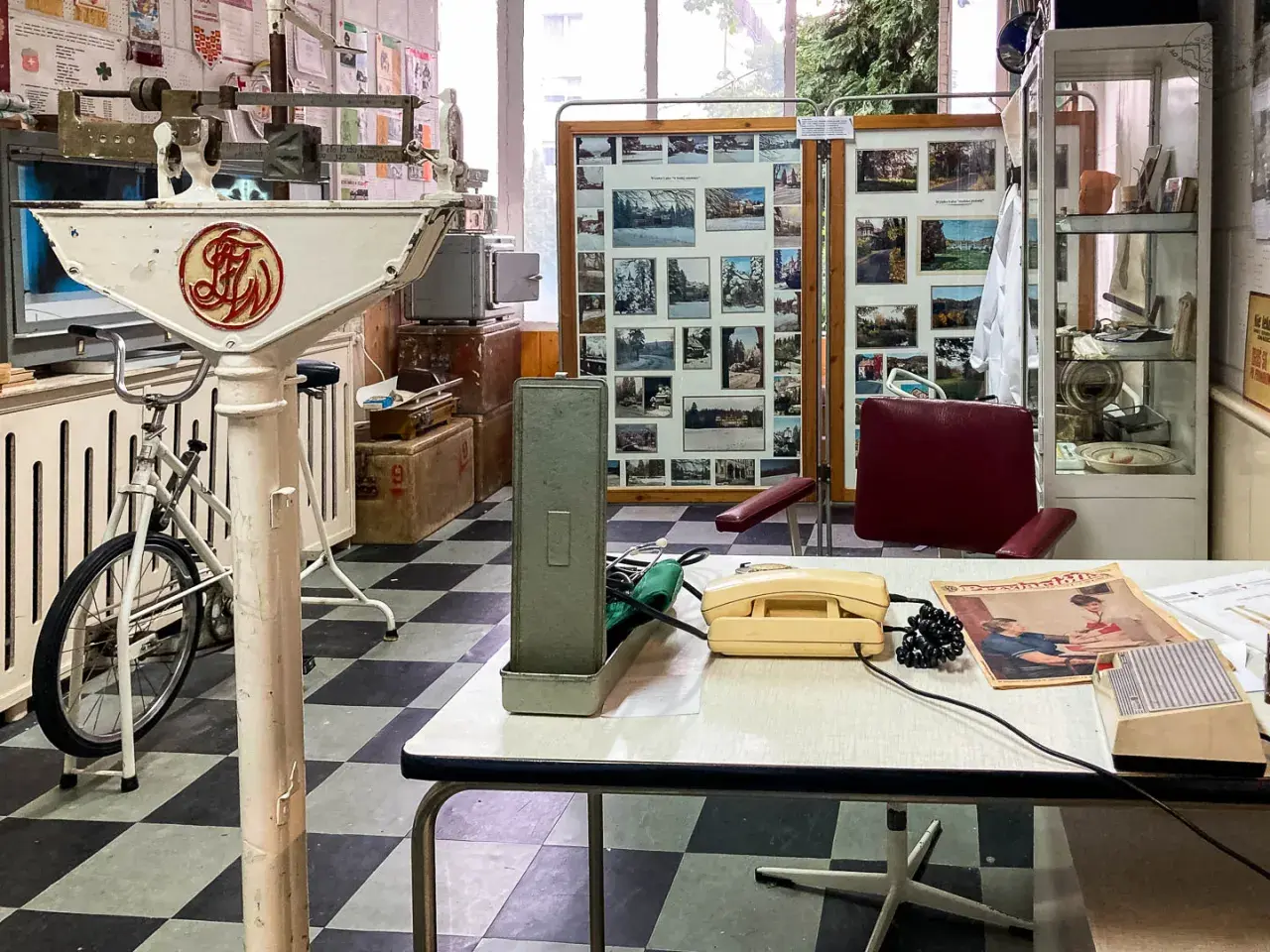 Wnętrze gabinetu lekarskiego z lat 70. XX wieku, część ekspozycji muzeum Dolny Śląsk. Widoczna waga lekarska, rower, aparat telefoniczny i zdjęcia.