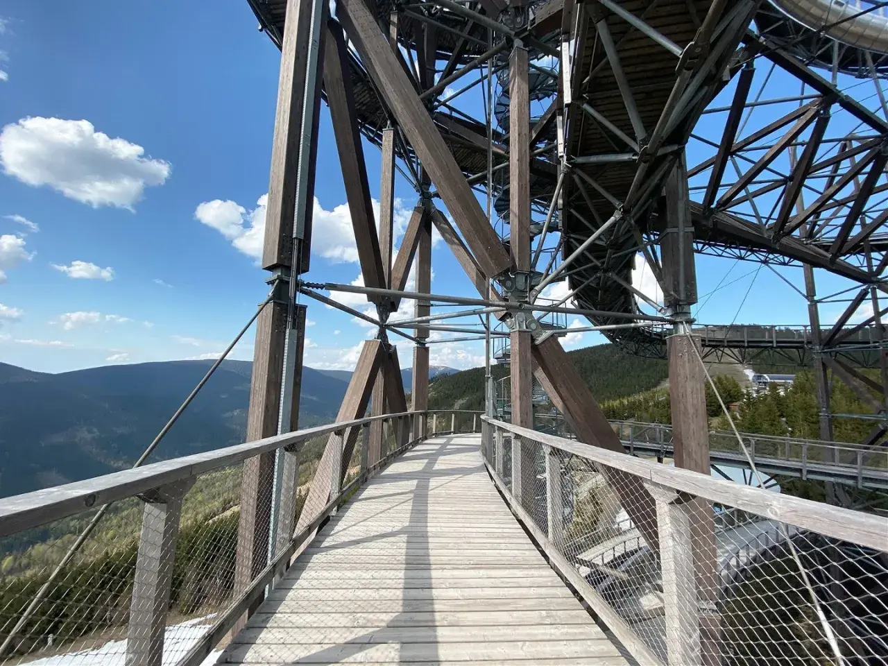 Drewniany sky bridge wznosi się nad górami, oferując widok na lasy i błękitne niebo z chmurami.