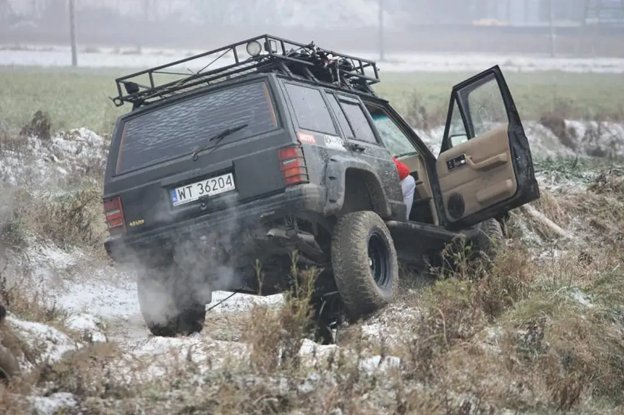 Czarny Jeep z bagażnikiem dachowym pokonuje trudny teren, to prawdziwe off road warszawa! Dym z opon i otwarta drzwi dodają dynamiki.