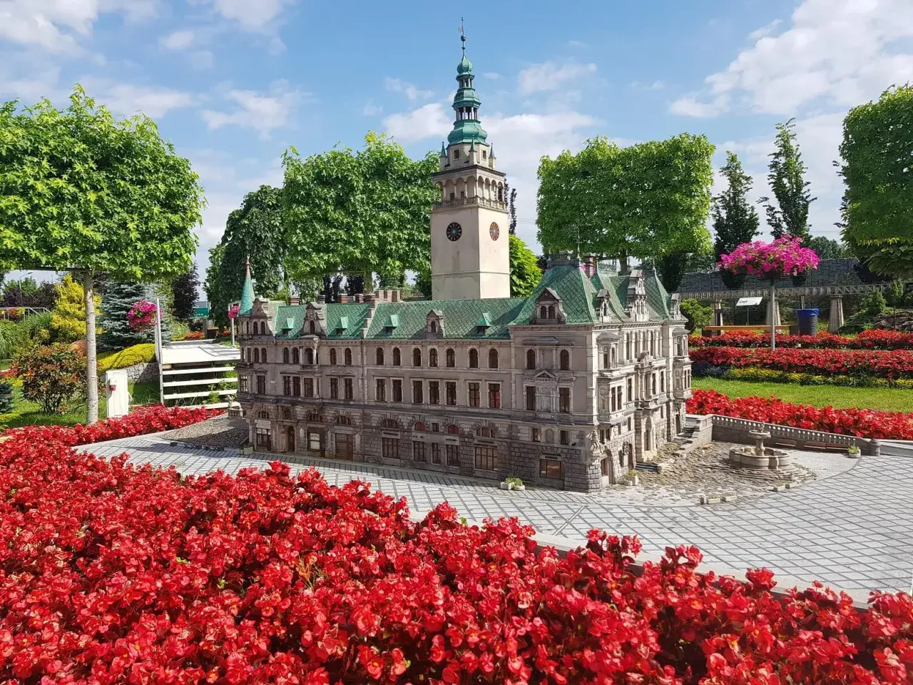 Miniatura ratusza z wieżą i zegarem, otoczona czerwonymi kwiatami. Jedna z wielu kłodzko atrakcje.