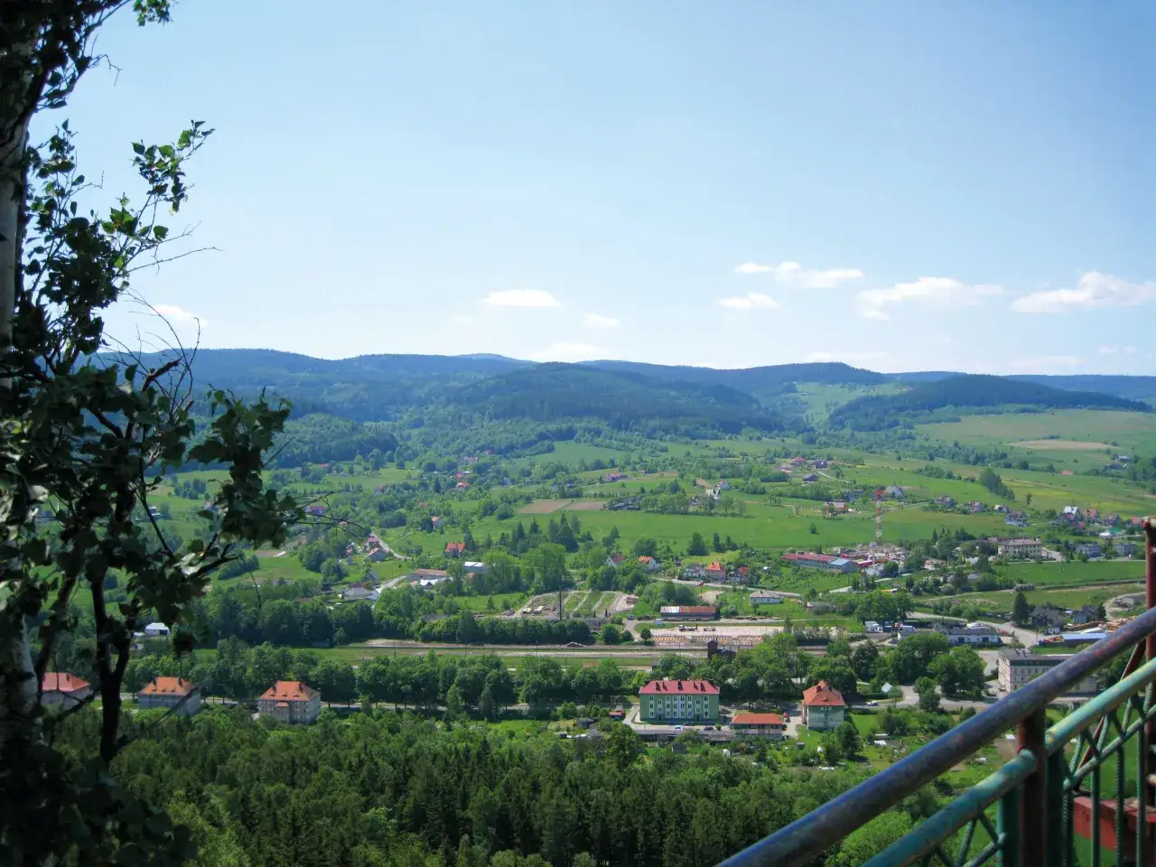 Widok na zieloną dolinę z zabudowaniami i lasami. Szczytna atrakcje turystyczne w oddali.