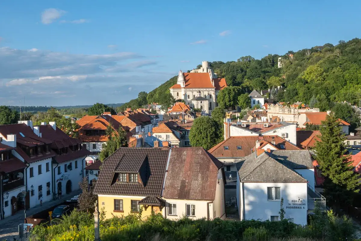 Widok na Kazimierz Dolny z dachem kościoła i wzgórzem. Widać urokliwe kamienice i zielone drzewa, tworzące malowniczy krajobraz. Kazimierz Dolny atrakcje zachwycają.