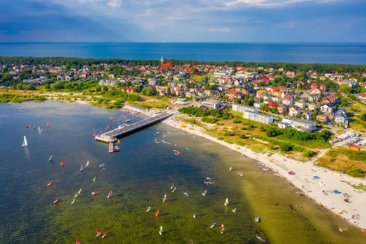 Jastarnia atrakcje: molo, plaża z turystami, zatoka pełna żeglarzy i windsurferów, w tle zabudowania i morze.