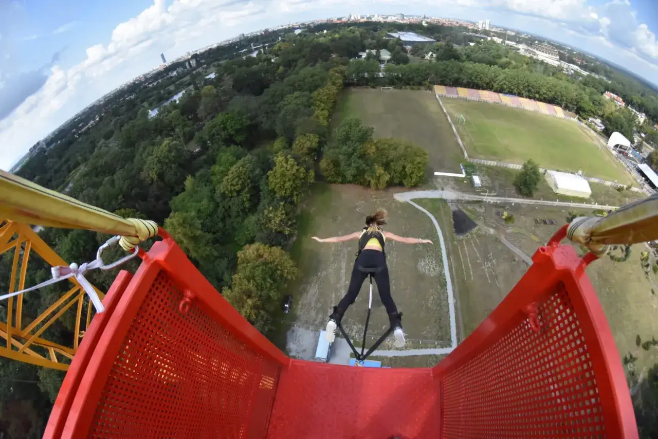 Kobieta skacze na bungee we Wrocławiu. Widok z g&oacute;ry na zielony park, boisko i miasto w oddali.