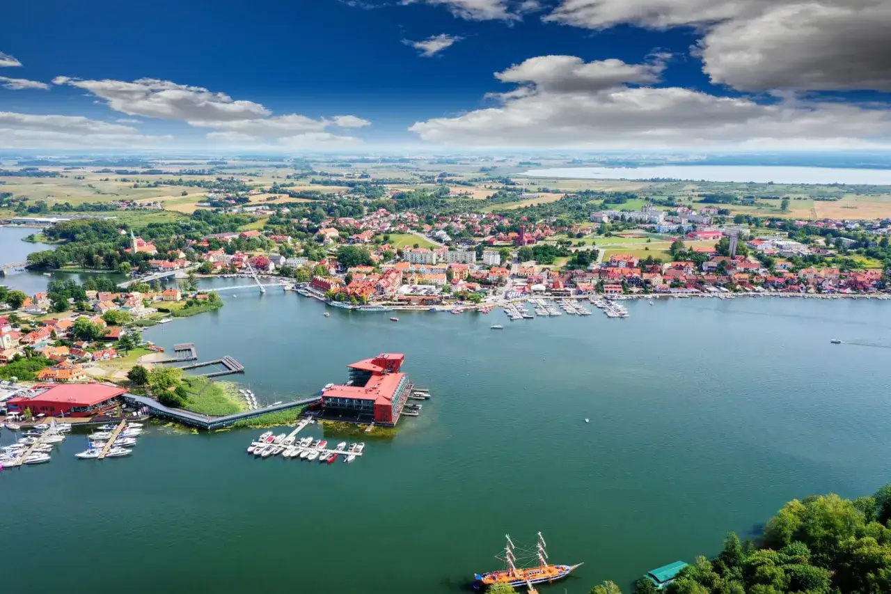 Malownicze mazury miejscowości nad jeziorem, z mariną pełną łodzi, czerwonym budynkiem i żaglowcem.