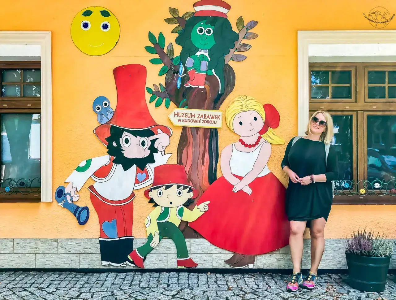 Kudowa-Zdr&oacute;j zaprasza do Muzeum Zabawek na Dolnym Śląsku! Kolorowa ściana z postaciami z bajek i uśmiechnięta kobieta.