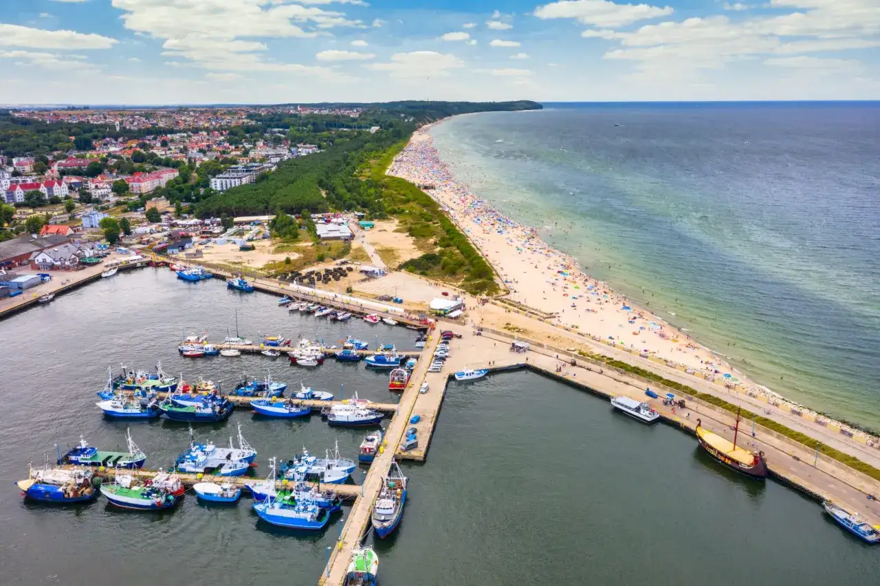 Port we Władysławowie i okolice tętnią życiem. Pełna łodzi zatoka, tłumy na plaży i zielone wzgórza tworzą malowniczy krajobraz.