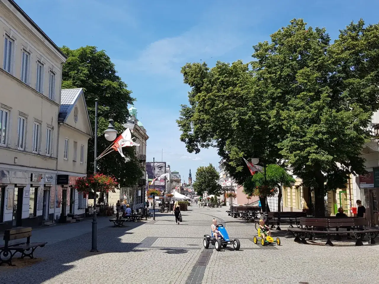 Dzieci ścigają się na pedałach po brukowanej ulicy w Radomiu. W tle widać kamienice, drzewa i polskie flagi, co tworzy urokliwy obraz miejskich atrakcji.