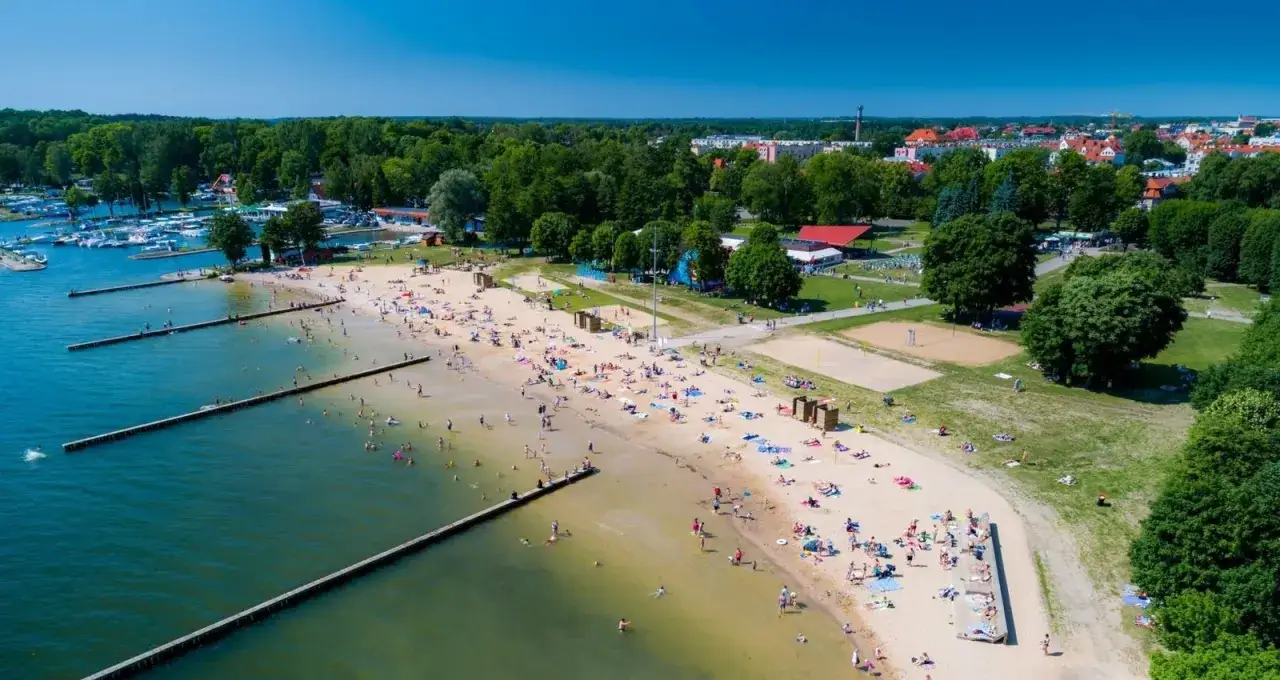 Tłoczna plaża na Mazurach, z ludźmi kąpiącymi się i opalającymi. W tle widać przystań z łodziami i zielone drzewa.