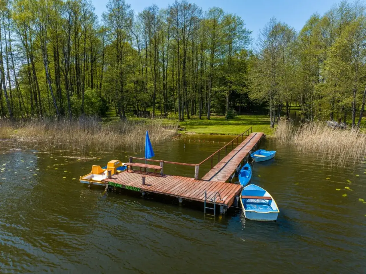 Idealne miejsce na wakacje nad wodą: pomost z ł&oacute;dkami i rowerkiem wodnym, otoczony lasem i trzcinami.