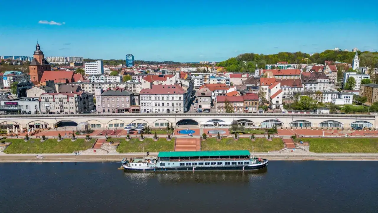 Widok na rzekę i miasto Gorzów Wielkopolski. Statek wycieczkowy cumuje przy nabrzeżu, a w tle widać zabytkową architekturę i tereny zielone. To jedne z wielu gorzów wielkopolski atrakcje.