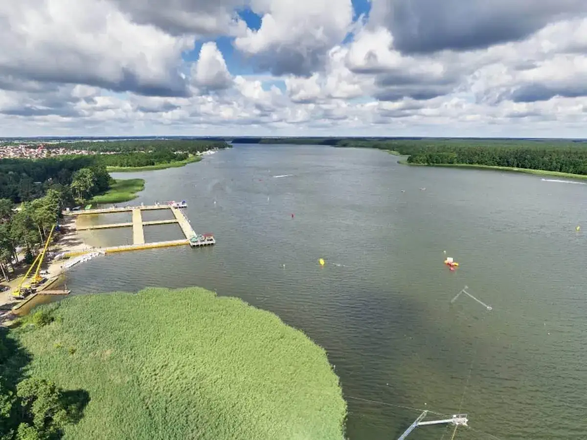 Malownicze jezioro z pomostami i plażą, idealne na letnie atrakcje w Augustowie. W oddali las i zabudowania.