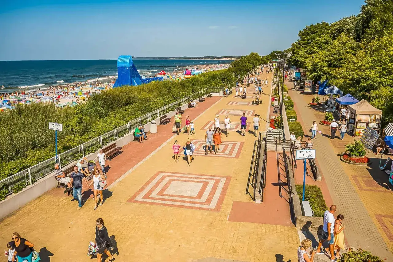 Promenada w Ustce tętni życiem. Ludzie spacerują, odpoczywają na ławkach, a w tle widać plażę i morze. To idealne miejsce na wakacyjne atrakcje.