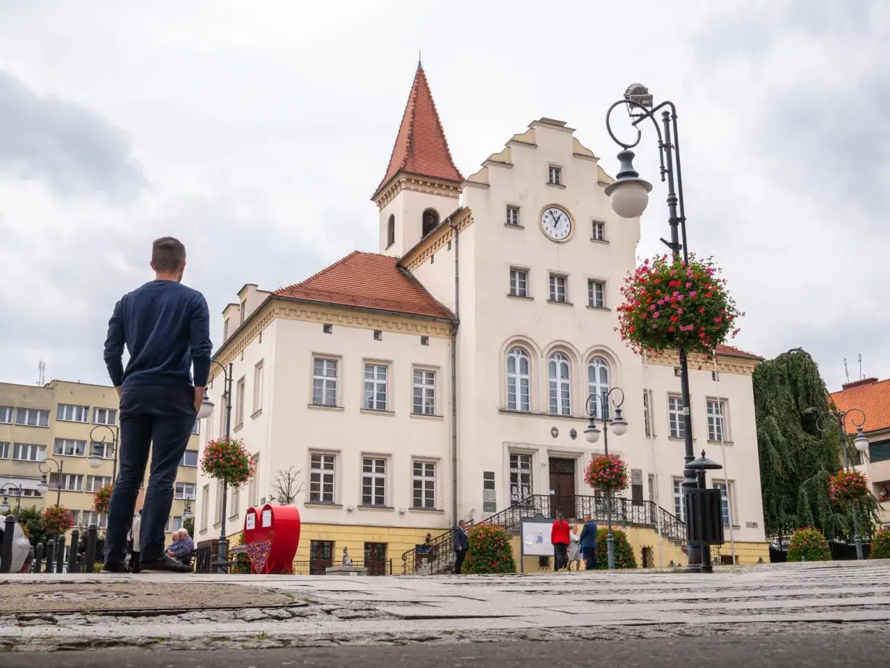 Mężczyzna podziwia ratusz w Trzebnicy, jedno z głównych atrakcji miasta. W tle zabytkowy budynek z zegarem i kwitnące pelargonie.