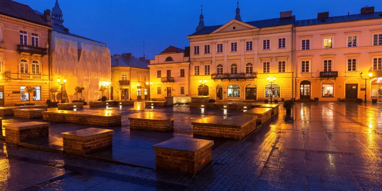 Piotrków Trybunalski atrakcje: Rynek o zmierzchu, mokry od deszczu, oświetlony latarniami i witrynami sklepów.