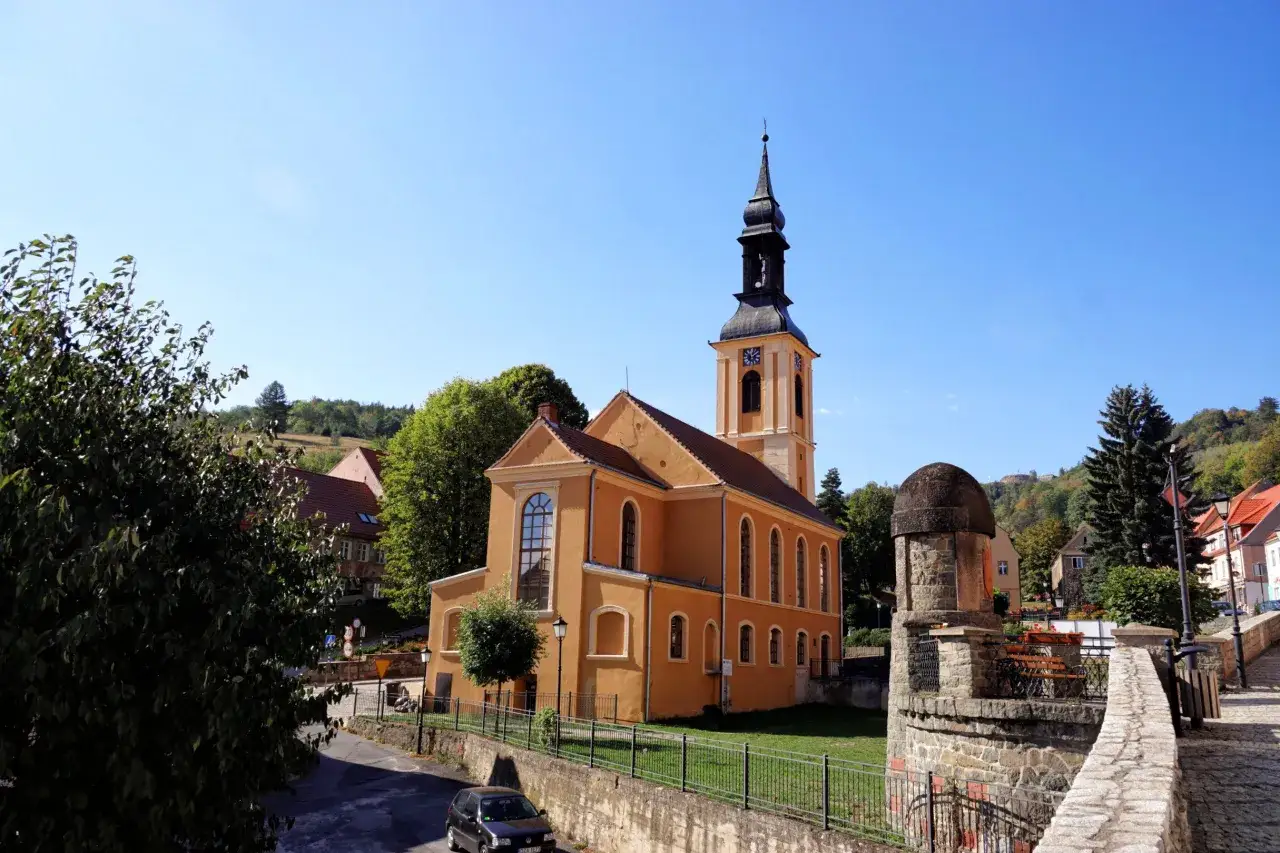 Pomarańczowy kości&oacute;ł z wieżą zegarową na rynku. W tle widać wzg&oacute;rza i zabudowania, a po prawej kamienną wieżyczkę.