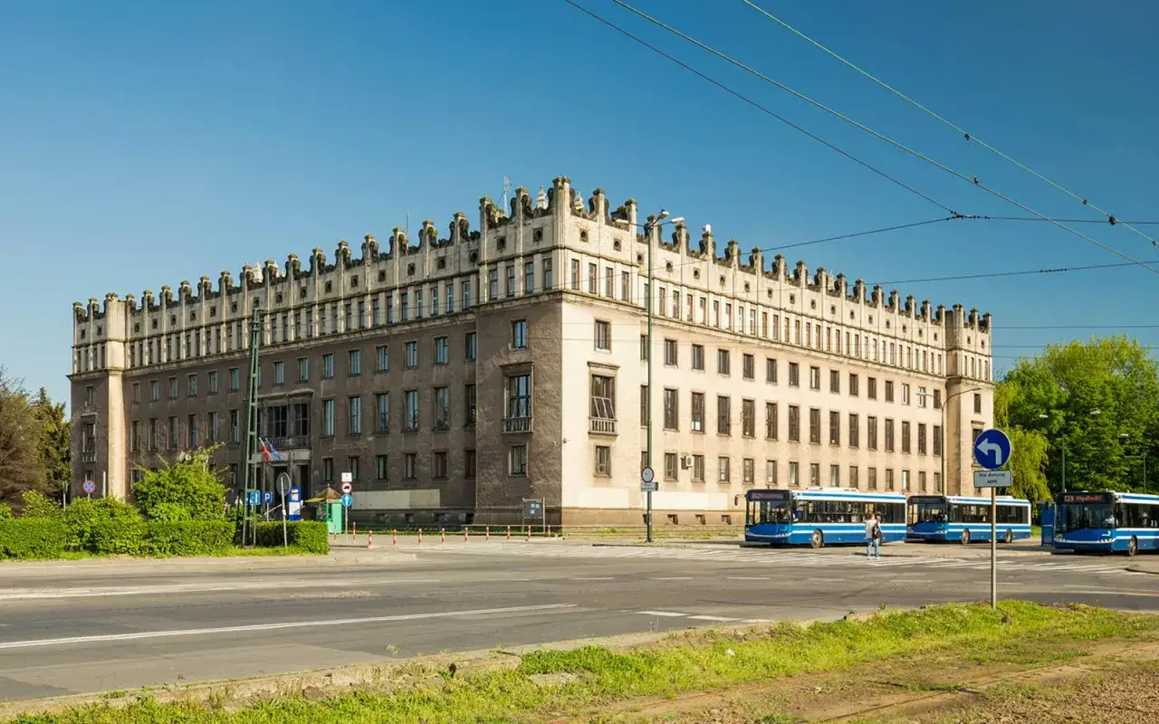 Imponujący budynek w stylu socrealistycznym, symbol Nowej Huty, z charakterystycznym fryzem i rzędami okien. Obok przejeżdżają niebieskie autobusy.