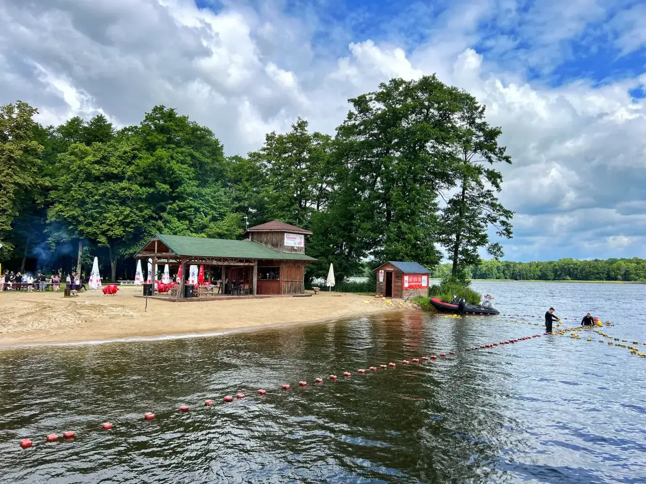 Plaża z barem i domkami nad jeziorem, idealne miejsce na letnie atrakcje w Szczecinku. Woda, piasek i zielone drzewa tworzą malowniczy krajobraz.