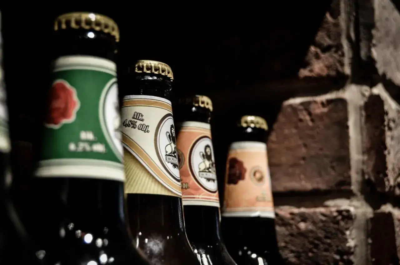 Butelki piwa w Poznaniu, gotowe na degustację. R&oacute;żne etykiety, złote kapsle, ceglana ściana w tle.