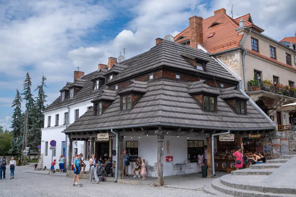 Urokliwy rynek w Kazimierzu Dolnym z charakterystycznymi kamieniczkami i drewnianymi dachami. Ludzie spacerują, kupują lody i pamiątki.