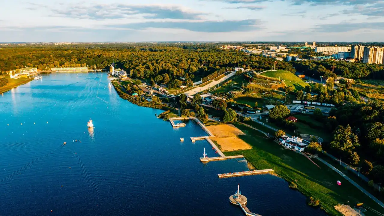 Widok z lotu ptaka na jezioro z pomostami, plażą i terenami zielonymi. W oddali las i zabudowania miejskie. To z pewnością fajne miejsca nad jeziorem.