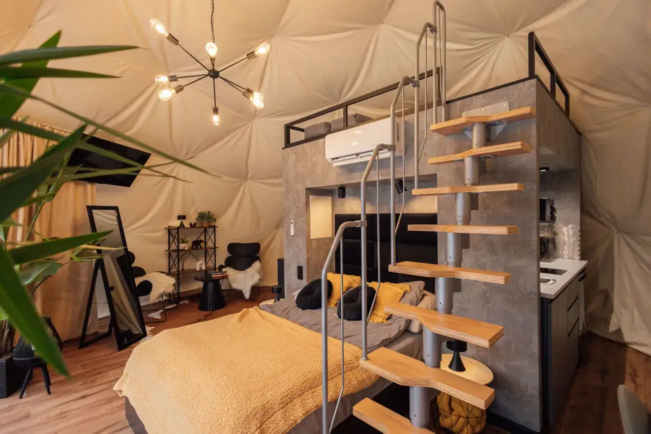 Nowoczesny glamping w Nałęczowie: przestronne wnętrze kopuły z ł&oacute;żkiem, schodami na antresolę i aneksem kuchennym.